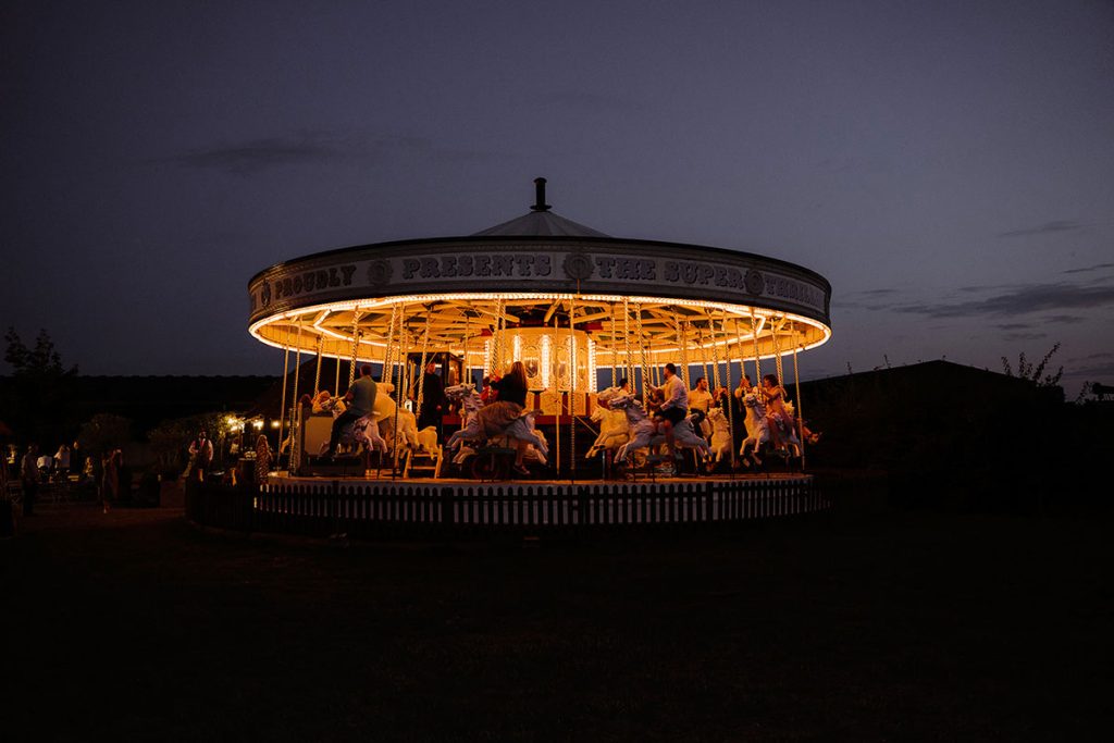 The carousel at Preston Court in Kent in the evening, lit up. A wedding group is visible on the carousel.