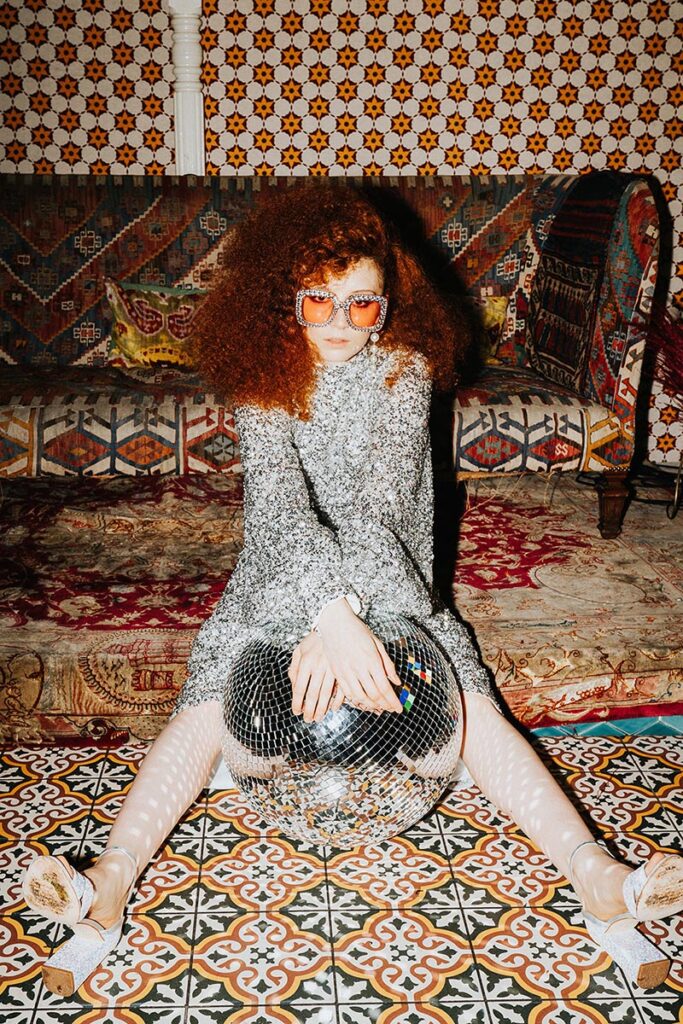 Editorial wedding portrait showing expressive movement and fashion-led styling in a maximalist setting with a bride in a silver sequin dress sitting on a patterned floor with a disco ball