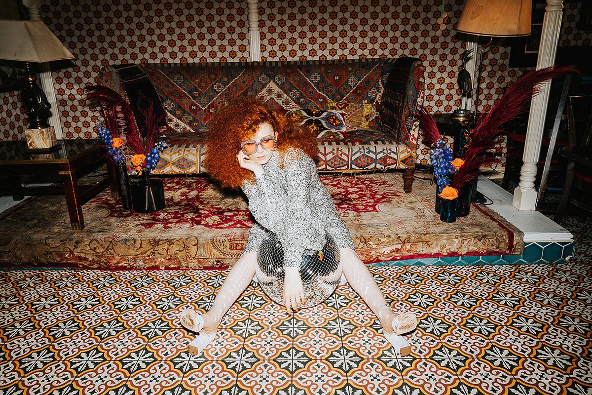 a bride at her maximalist wedding sitting on a tiled floor in a silver sequin dress holding a disco ball