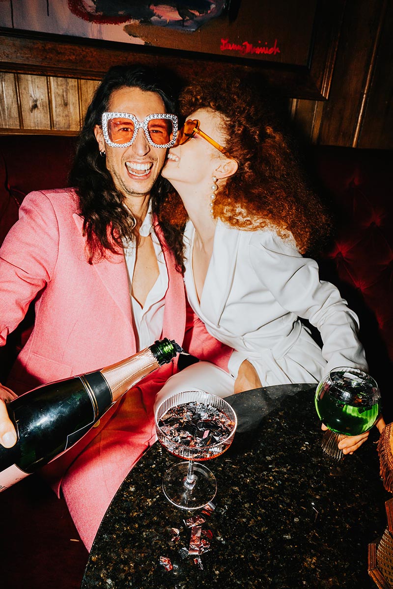 A couple celebrating their maximalist wedding by pouring champagne. The groom is dressed in a pink suit with embellished sunglasses, the bride is kissing his cheek and wearing a white suit