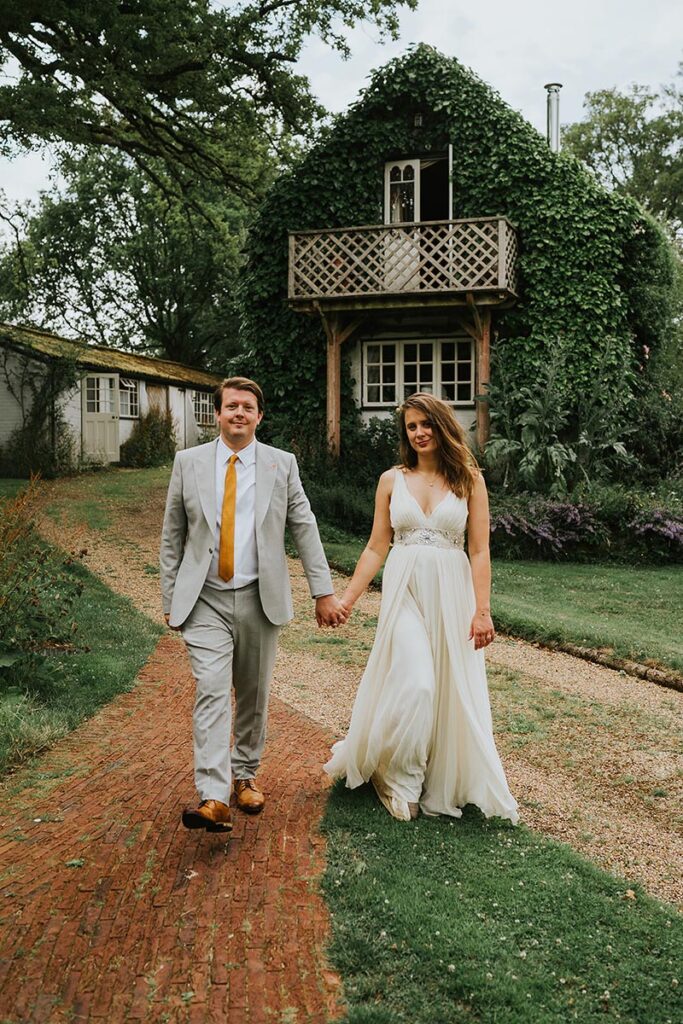 Couple at their Ridge Farm wedding waling away from a small building with a balcony covered in ivy.