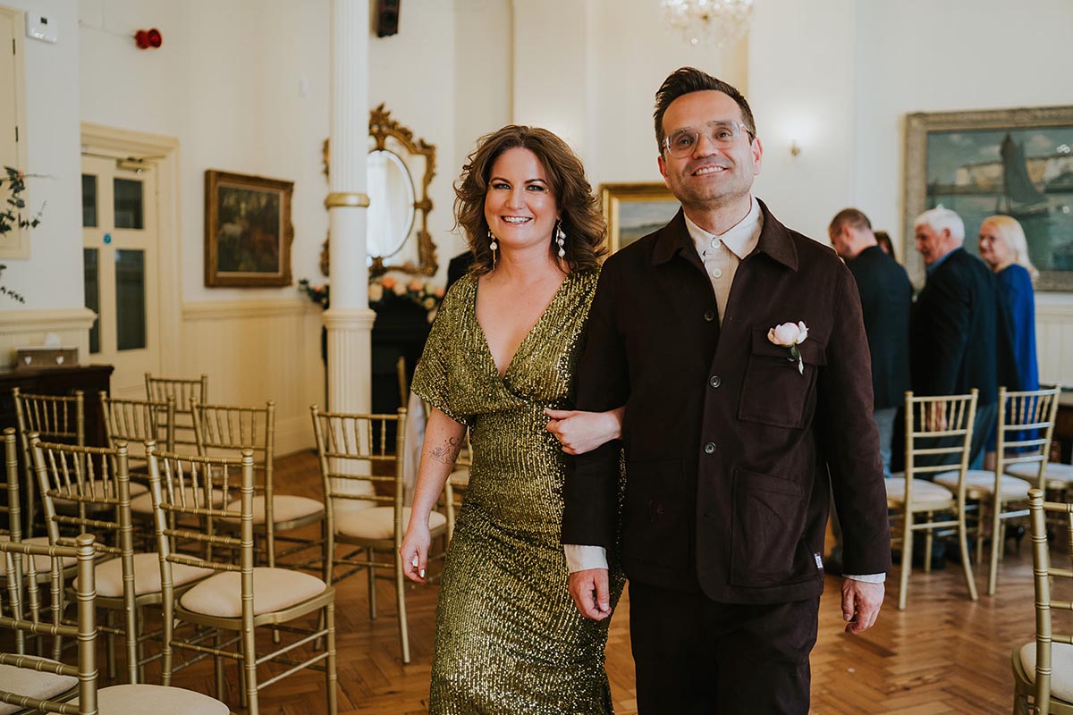 A couple exit their wedding at brighton town hall, the bride wears a green sequin dress and the groom wears a burgundy suit, they have linked arms and smile at the camera