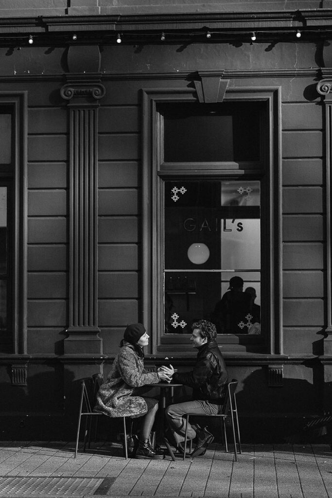 A couple sit at a table on the pavement outside a coffee shop for this cinematic engagement photography
