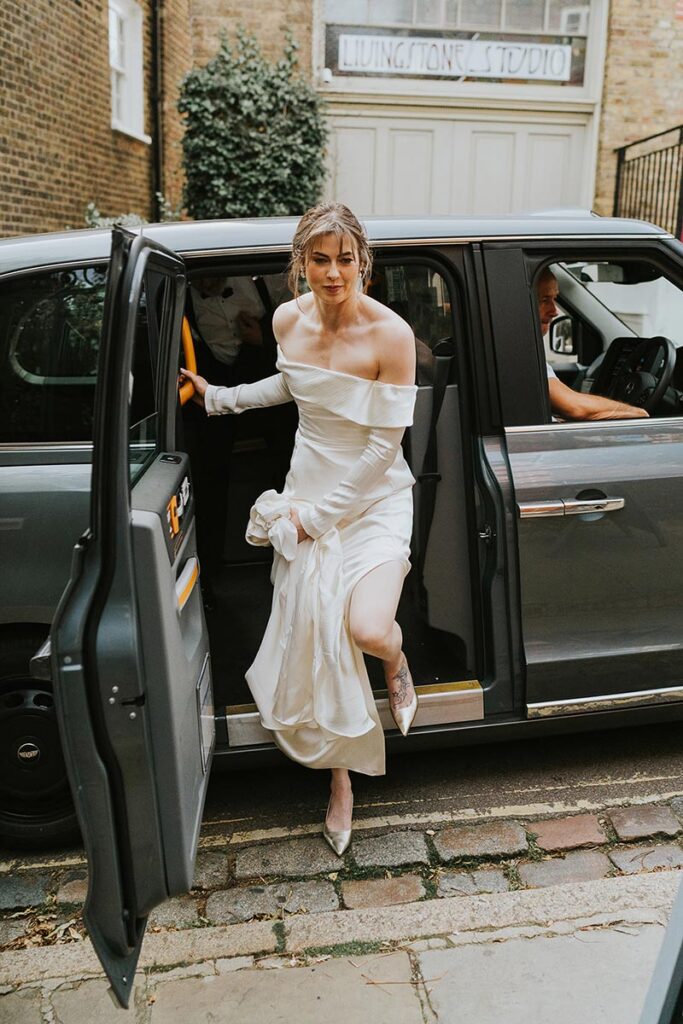 Bride in off-shoulder silk wedding dress stepping out of a London black cab, editorial wedding photography London