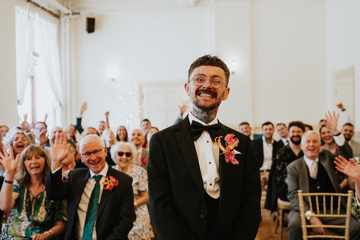 groom smiles at the camera as his guests wave behind him before his bride arrives for his brighton town hall wedding