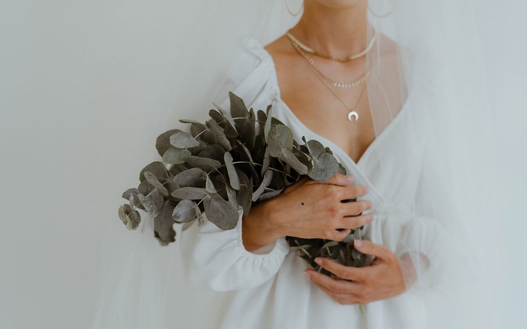 Picture of a bride holding a bunch of eucalyptus to her chest. The frame is cropped at her chin and waist, she wears a veil and gold necklaces and it is against a white background.