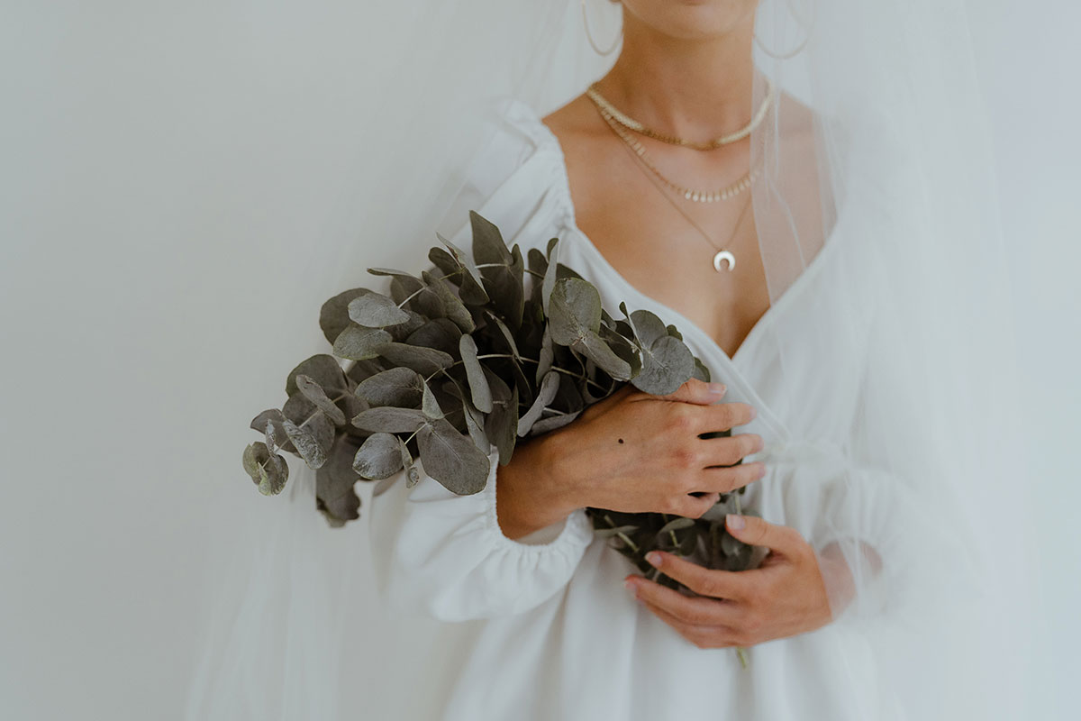 Picture of a bride holding a bunch of eucalyptus to her chest. The frame is cropped at her chin and waist, she wears a veil and gold necklaces and it is against a white background.