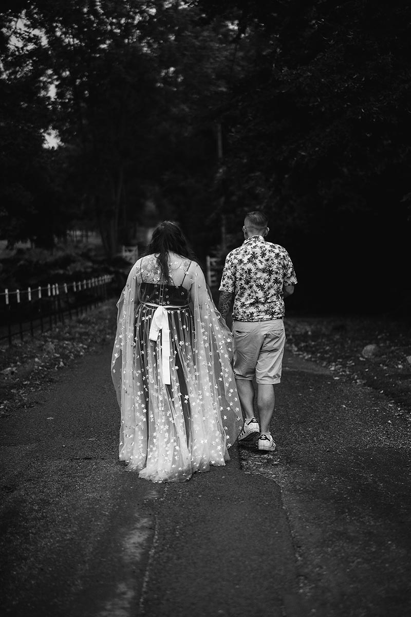 A wedding couple walk away from the camera at their wedding venue, Preston Court in Kent. The bride wears a cape covered in stars and the groom wears an Hawaiian shirt and shorts.